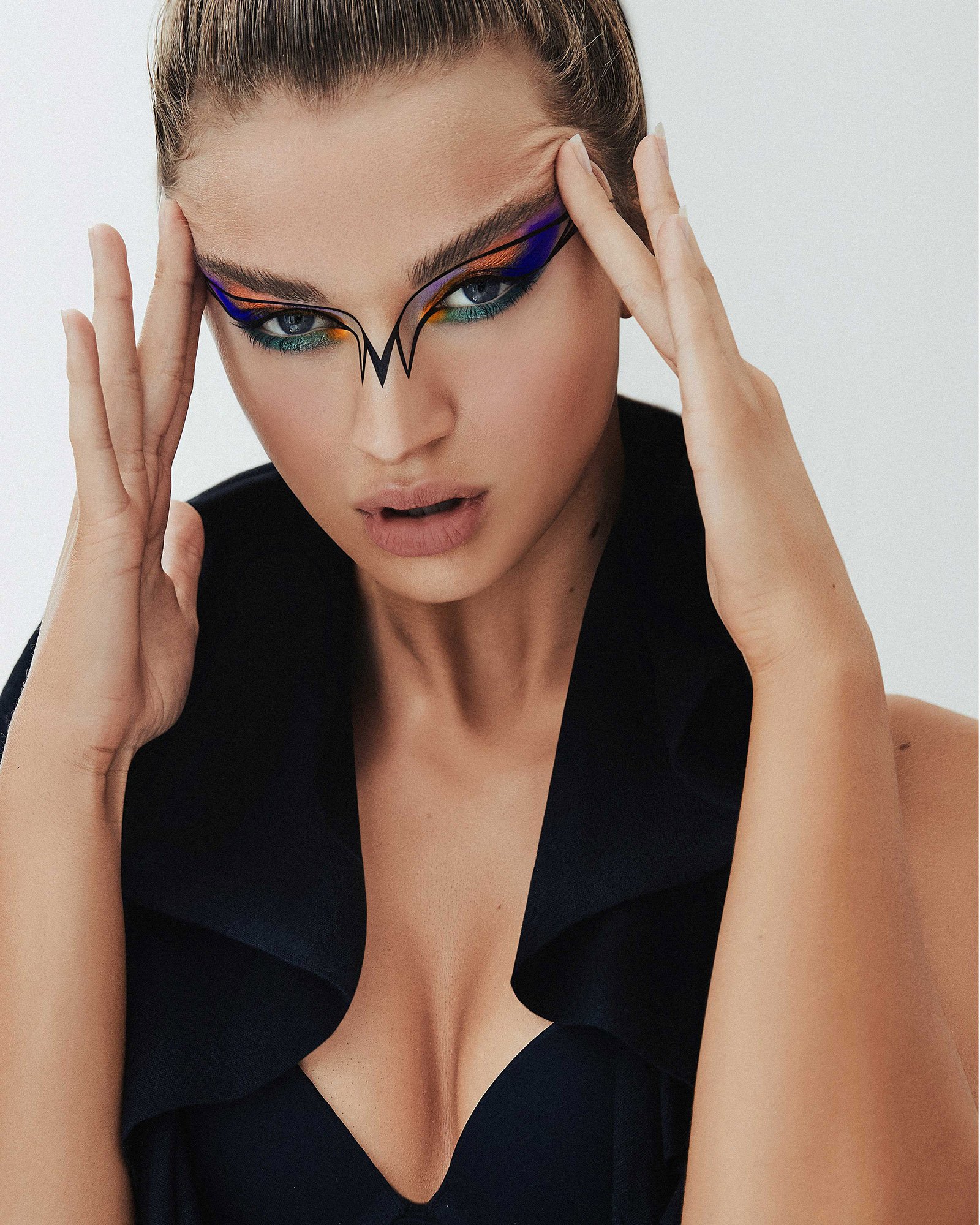 Portrait of a model with blue eyes and creative geometric makeup, her hands raised to her temples against a pale backdrop.