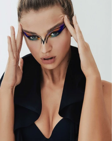 Portrait of a model with blue eyes and creative geometric makeup, her hands raised to her temples against a pale backdrop.