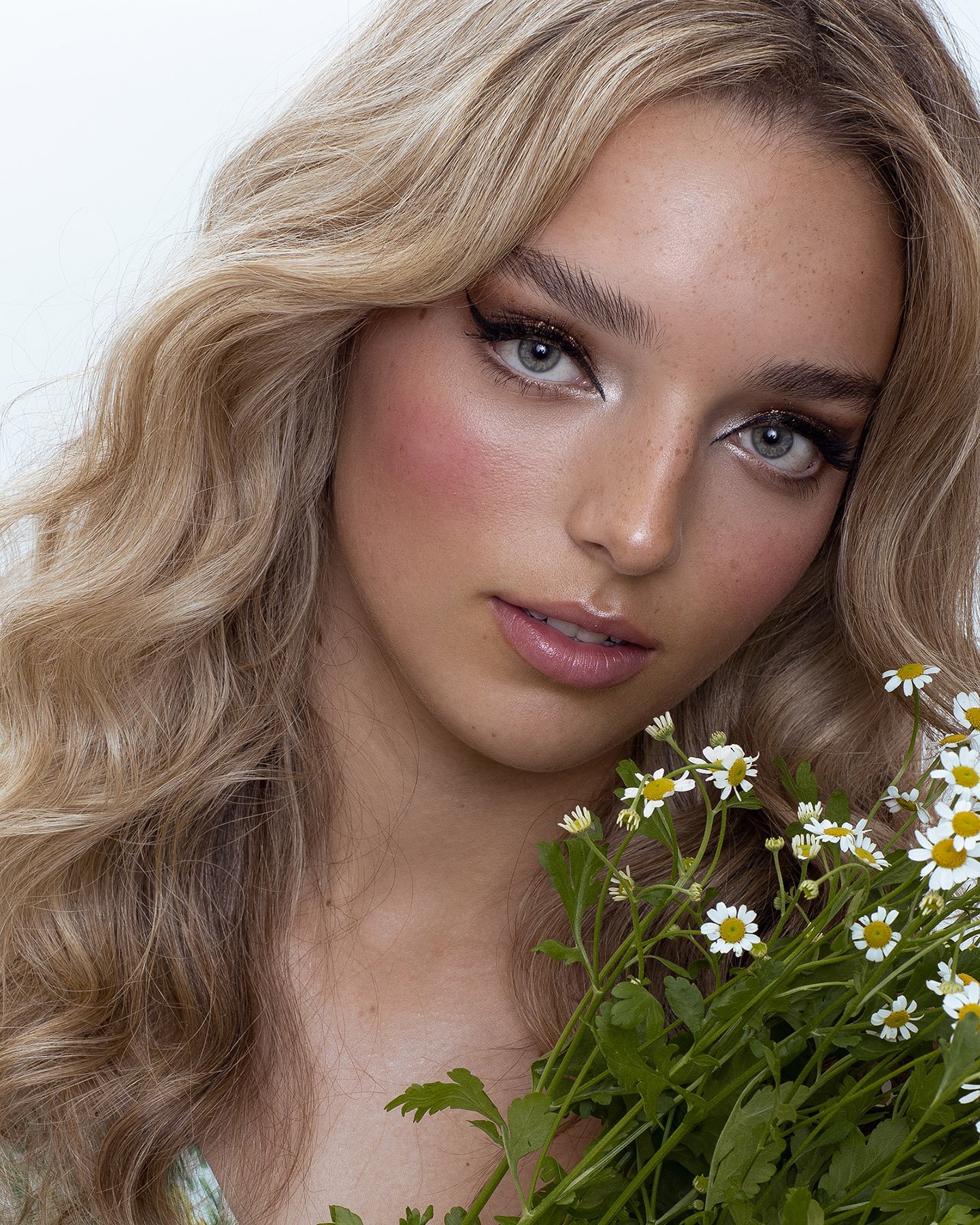 Close-up portrait of a blonde woman holding a bouquet of daisies against a clean white background in a beauty or fashion context.