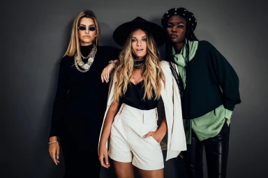 Three female models posing against a grey backdrop with styled hair and fashionable contemporary clothing