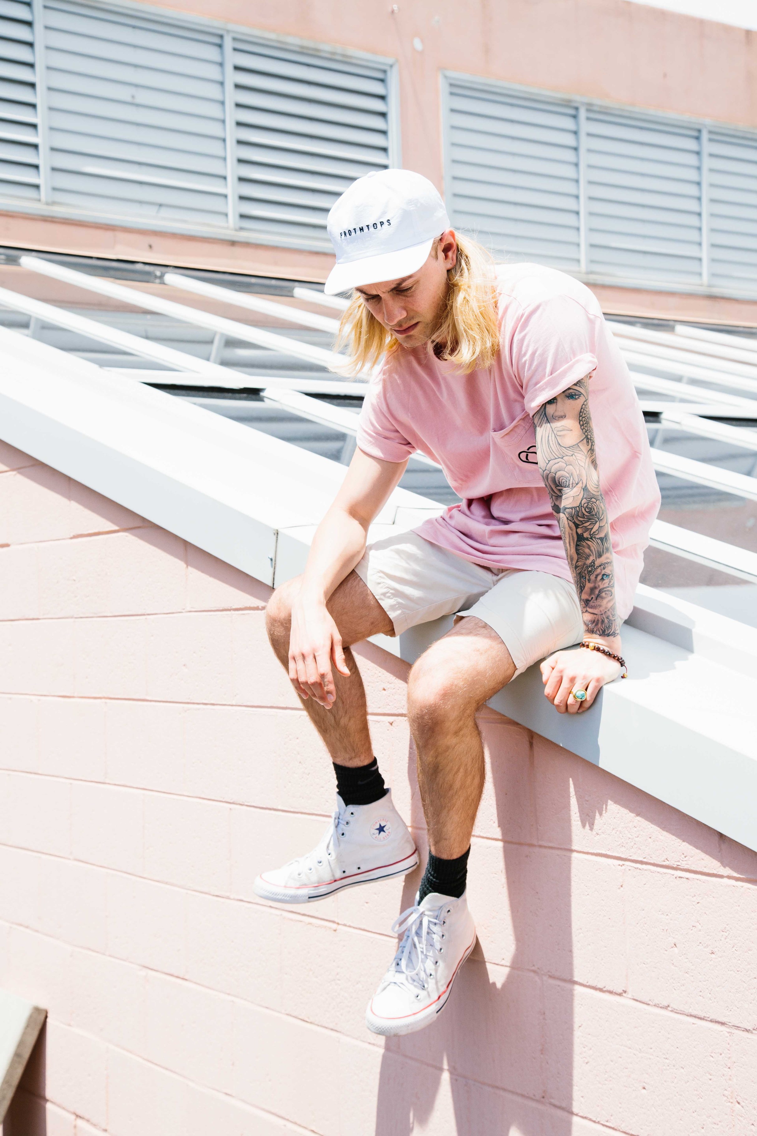 Young adult sits on pink roof ledge with light-pink shirt, cap, and sneakers. Blonde long hair with dark socks and arm tattoo.