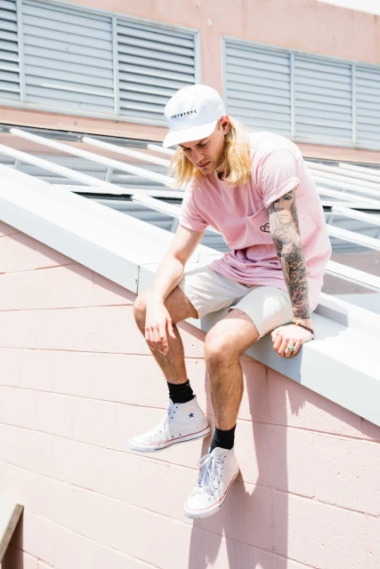Young adult sits on pink roof ledge with light-pink shirt, cap, and sneakers. Blonde long hair with dark socks and arm tattoo.