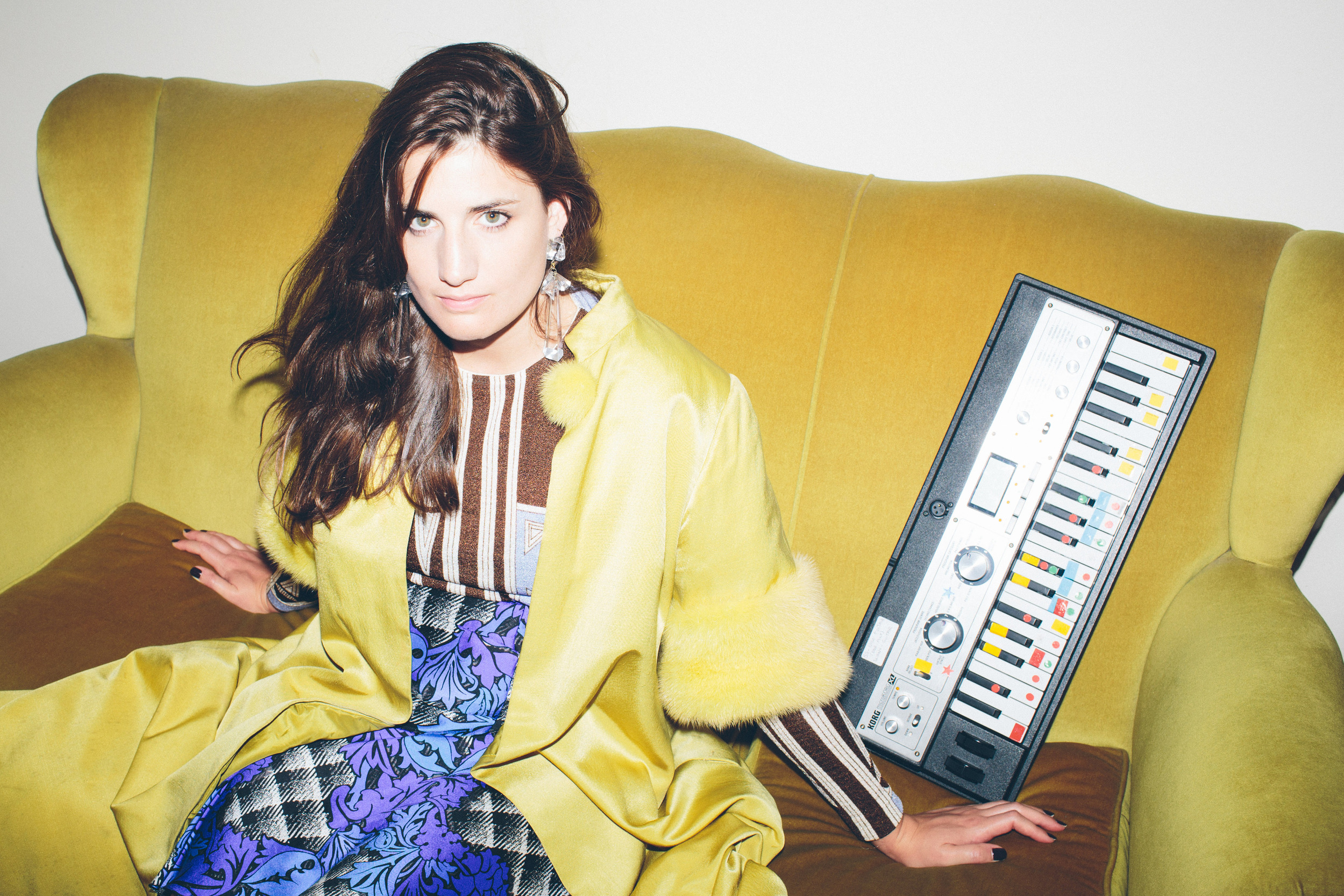 Portrait of a young adult woman sitting on a yellow velvet couch next to a keyboard with colorful buttons in an indoor setting.