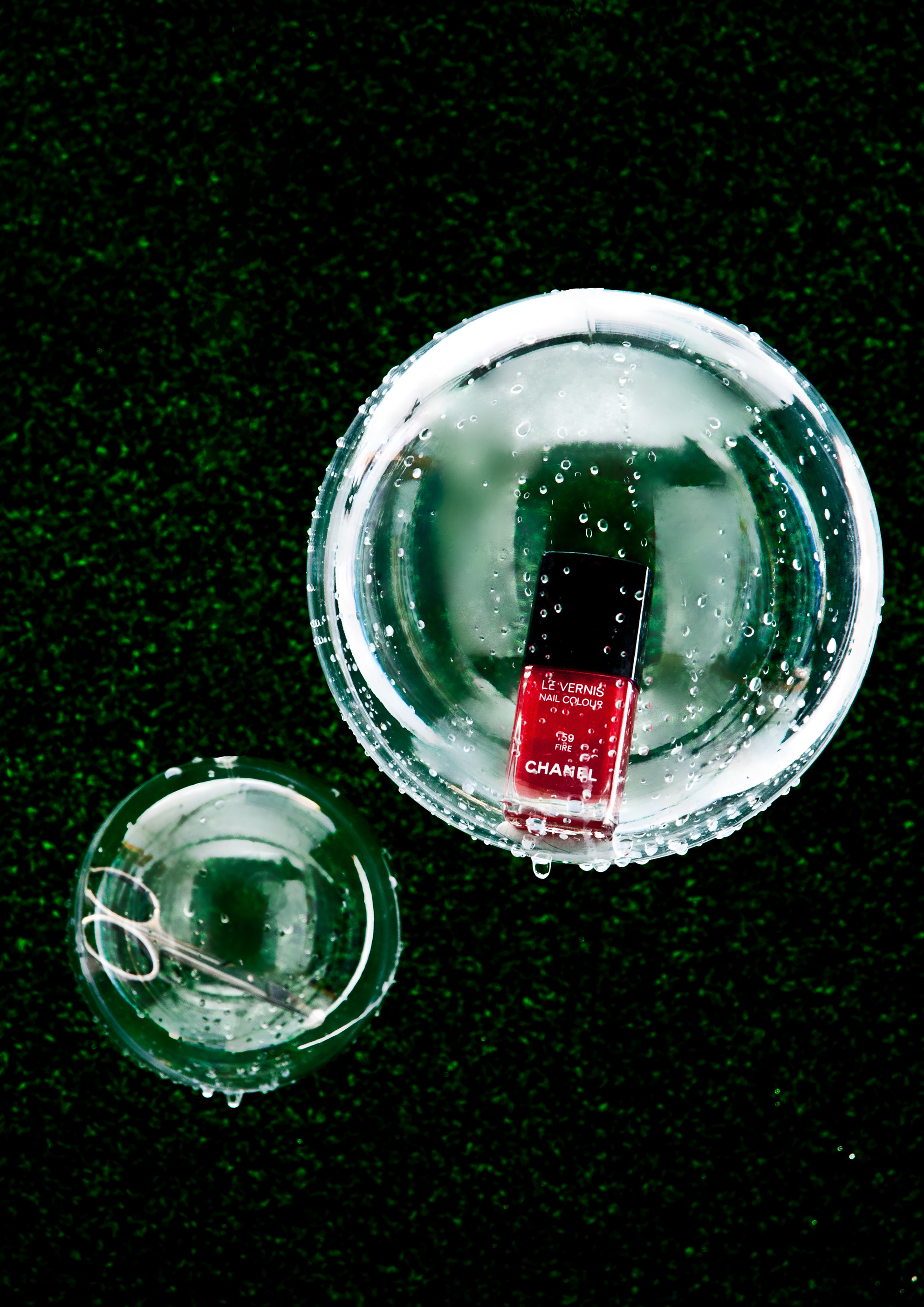 Overhead shot of a red nail polish bottle and scissors inside glass bowls on green textured turf