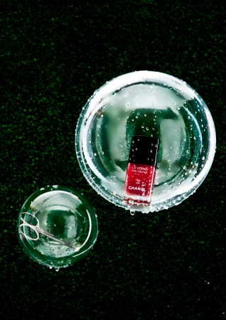 Overhead shot of a red nail polish bottle and scissors inside glass bowls on green textured turf