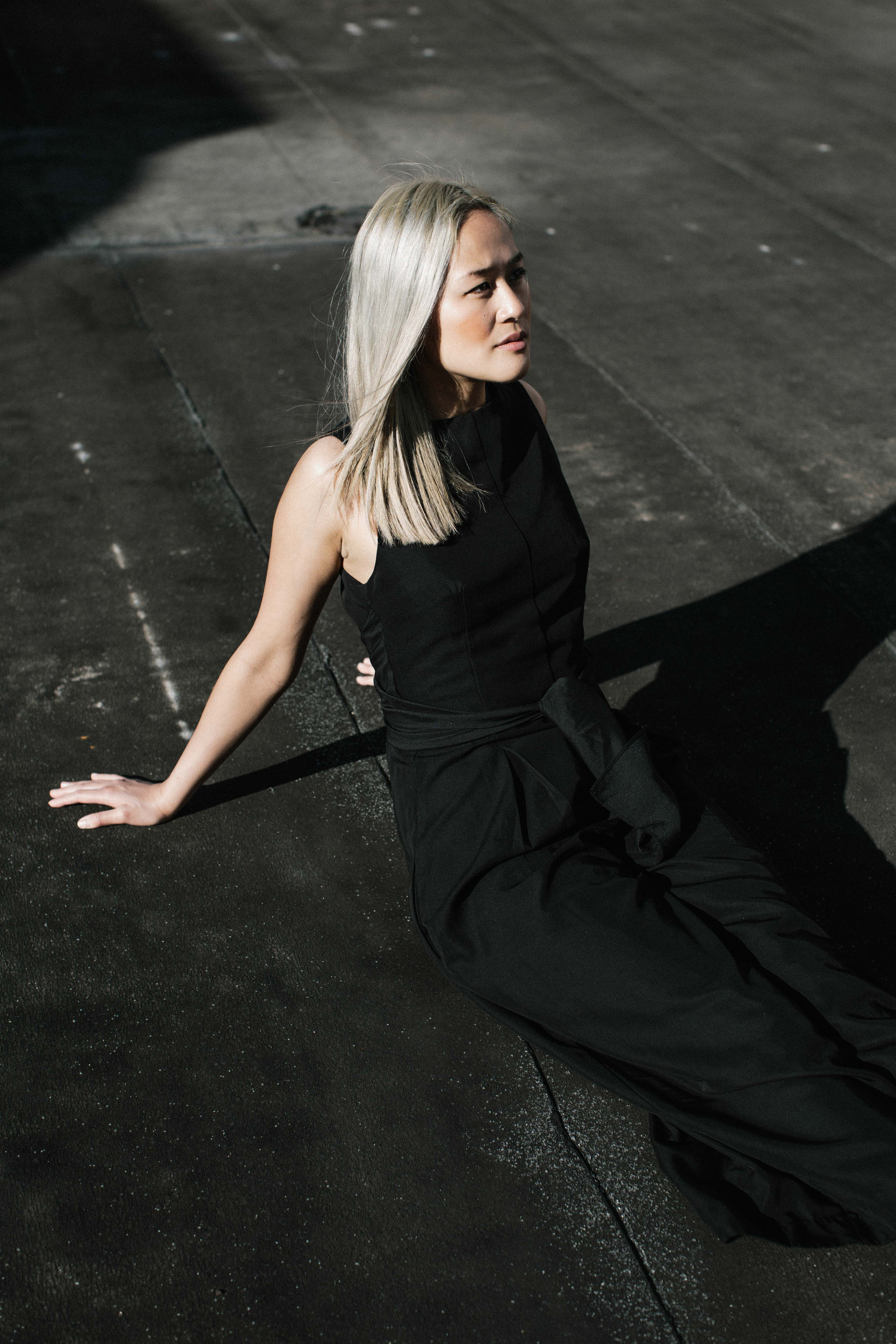 Woman with platinum blonde hair wearing a black sleeveless top and black pants, sitting on a black surface in direct sunlight.