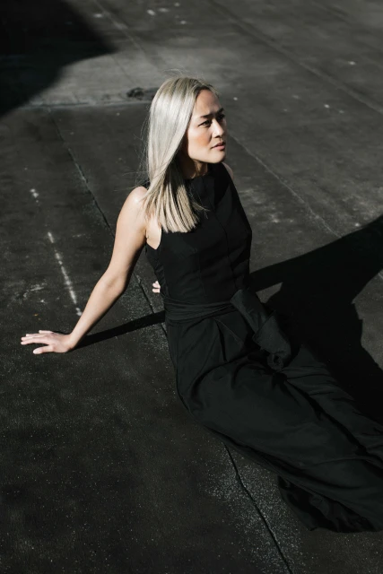 Woman with platinum blonde hair wearing a black sleeveless top and black pants, sitting on a black surface in direct sunlight.