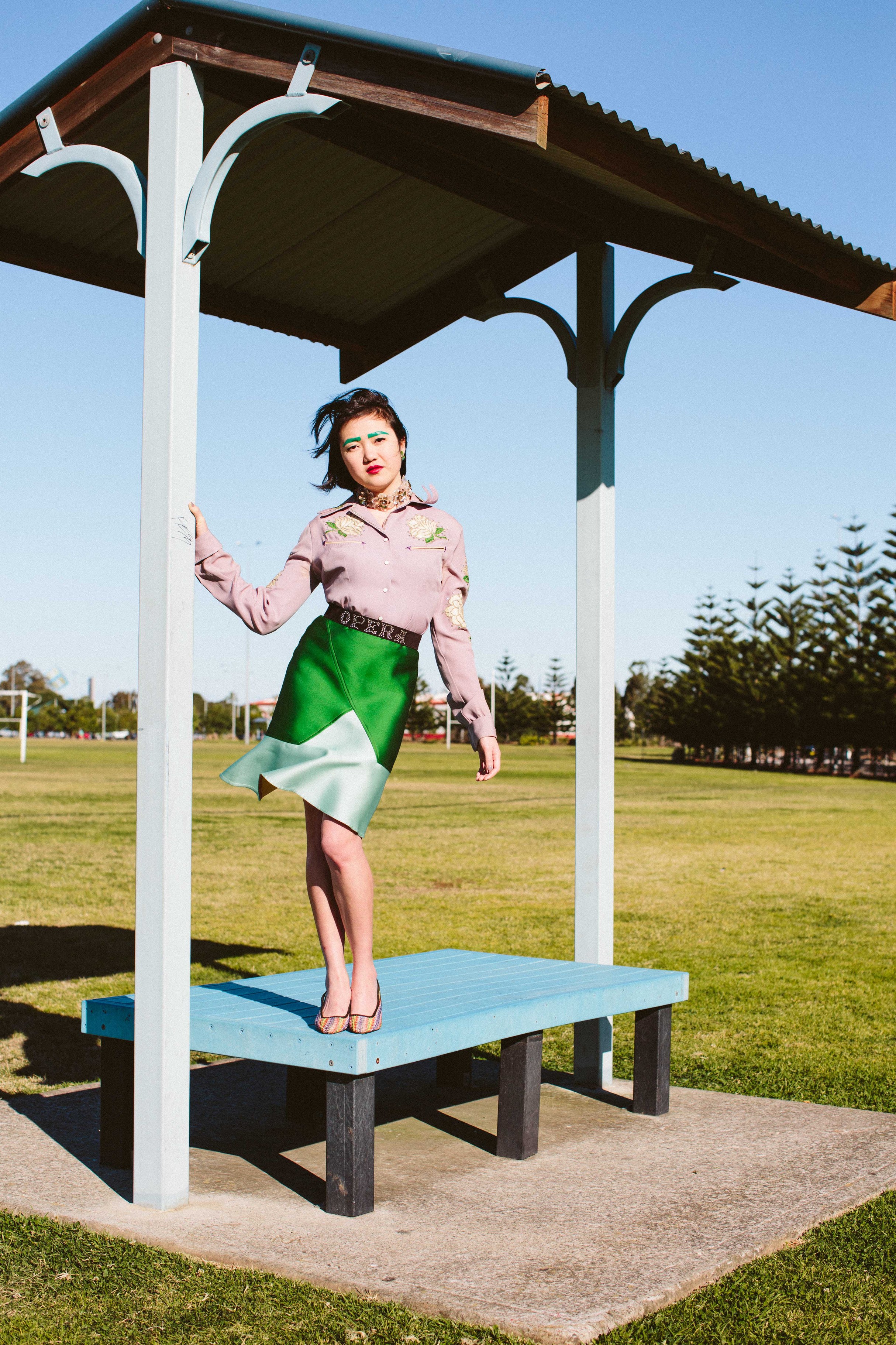 A woman poses in a stylized green skirt and mauve blouse under a blue outdoor structure in a park with trees and grass under sunny sky.
