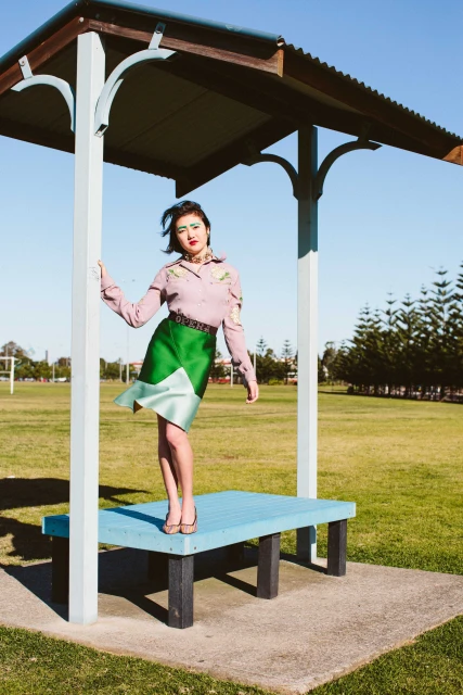 A woman poses in a stylized green skirt and mauve blouse under a blue outdoor structure in a park with trees and grass under sunny sky.