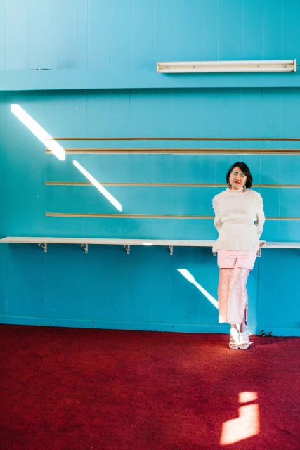 A woman in a white sweater and pink skirt stands in a blue room with a red carpet, illuminated by sunlight and hanging shelves.