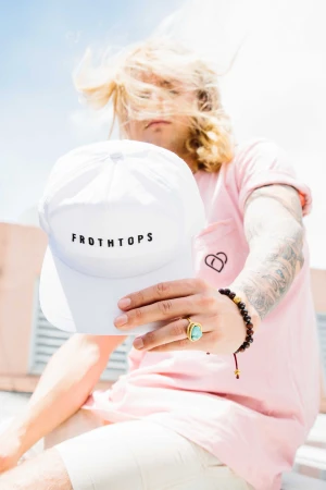 Man holding a white Frothtops baseball cap toward the camera while wearing a pink t-shirt in Sydney.