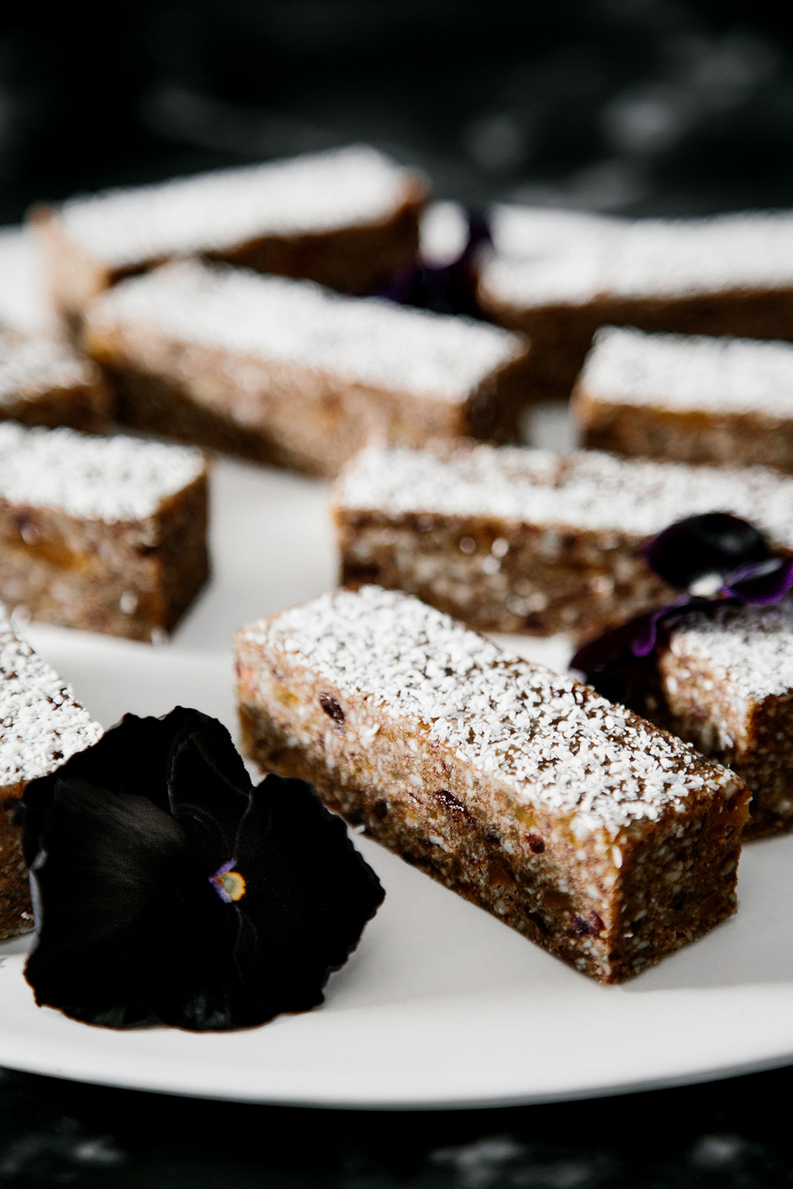 Close-up of rectangular desserts dusted with powdered sugar on a white plate, accented by dark purple flower petals against black backdrop.