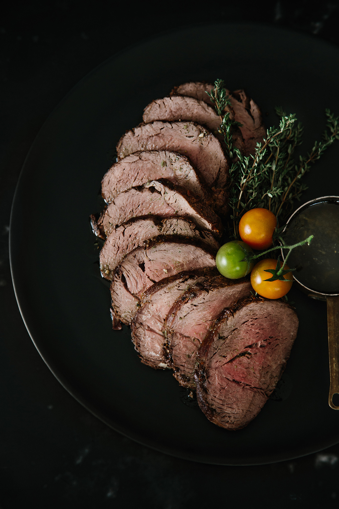 Overhead shot of sliced beef tenderloin on a black plate with fresh thyme sprigs and cherry tomatoes, paired with a side of gravy.