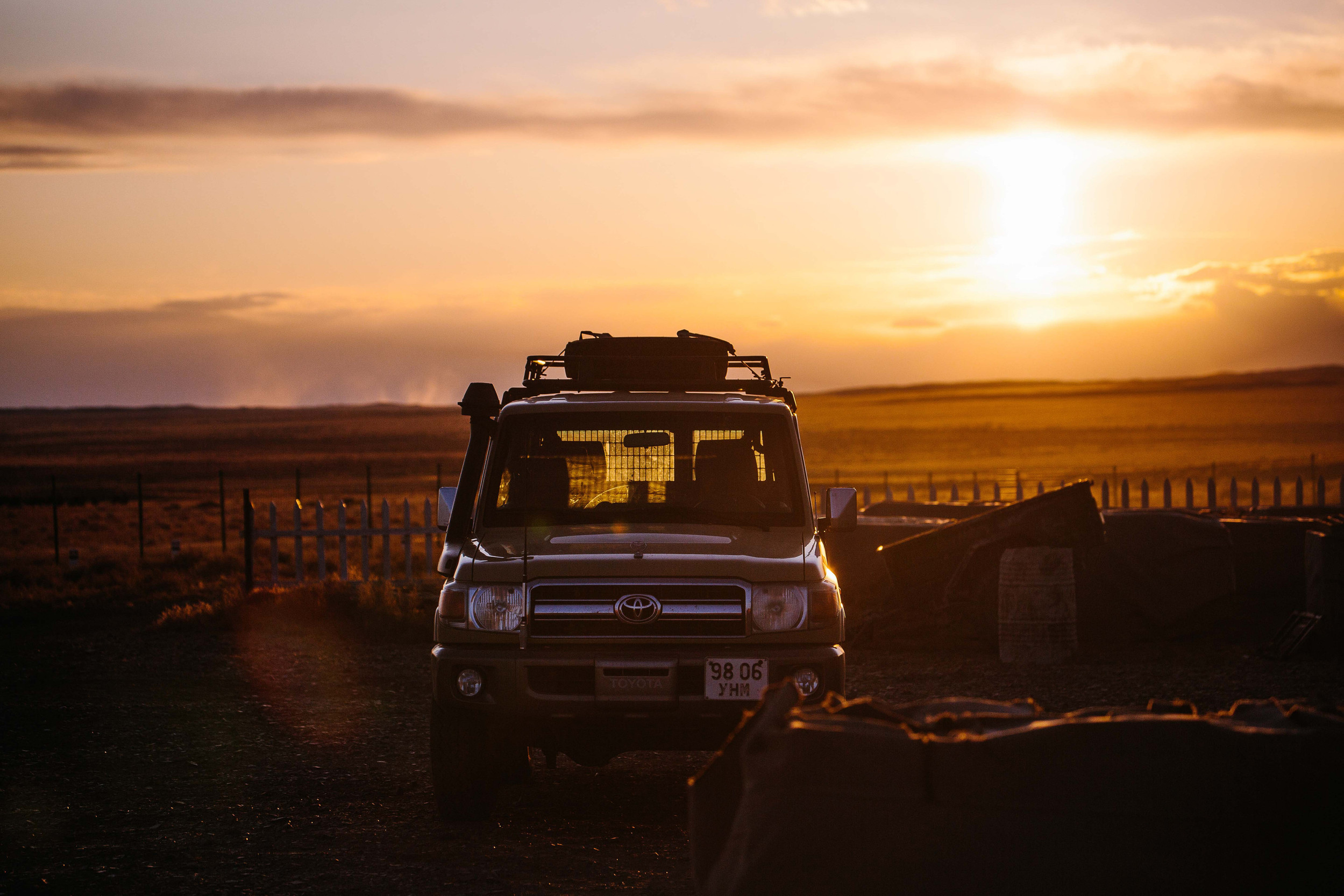 An off-road vehicle is parked in a rural setting during a golden sunset, with a landscape of fields and a partially visible white fence.