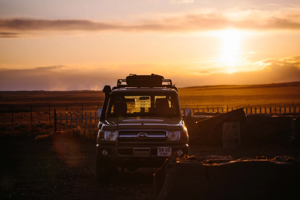 An off-road vehicle is parked in a rural setting during a golden sunset, with a landscape of fields and a partially visible white fence.