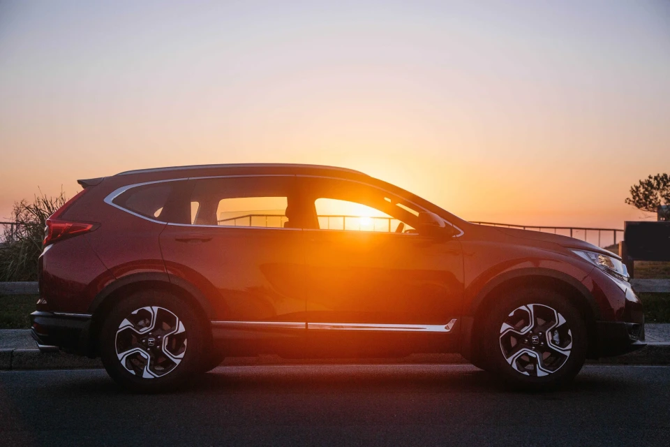 Red SUV parked on asphalt at golden hour, sun reflecting in windows. Side view of car with rims, tires, and body details.