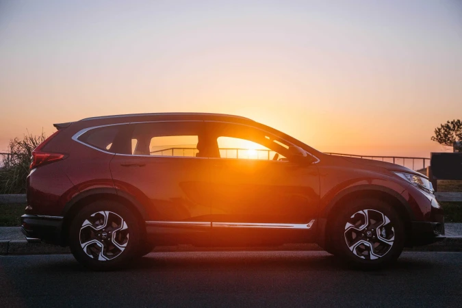 Red SUV parked on asphalt at golden hour, sun reflecting in windows. Side view of car with rims, tires, and body details.