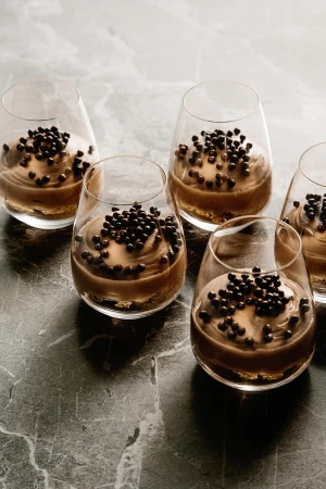Several clear glasses filled with a layered chocolate dessert, topped with dark brown sprinkles, arranged on a gray marble surface.
