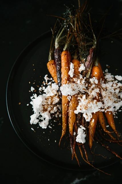 Overhead shot of roasted carrots covered in white crumbles on a black plate, food photography, minimalist composition, dark and moody.