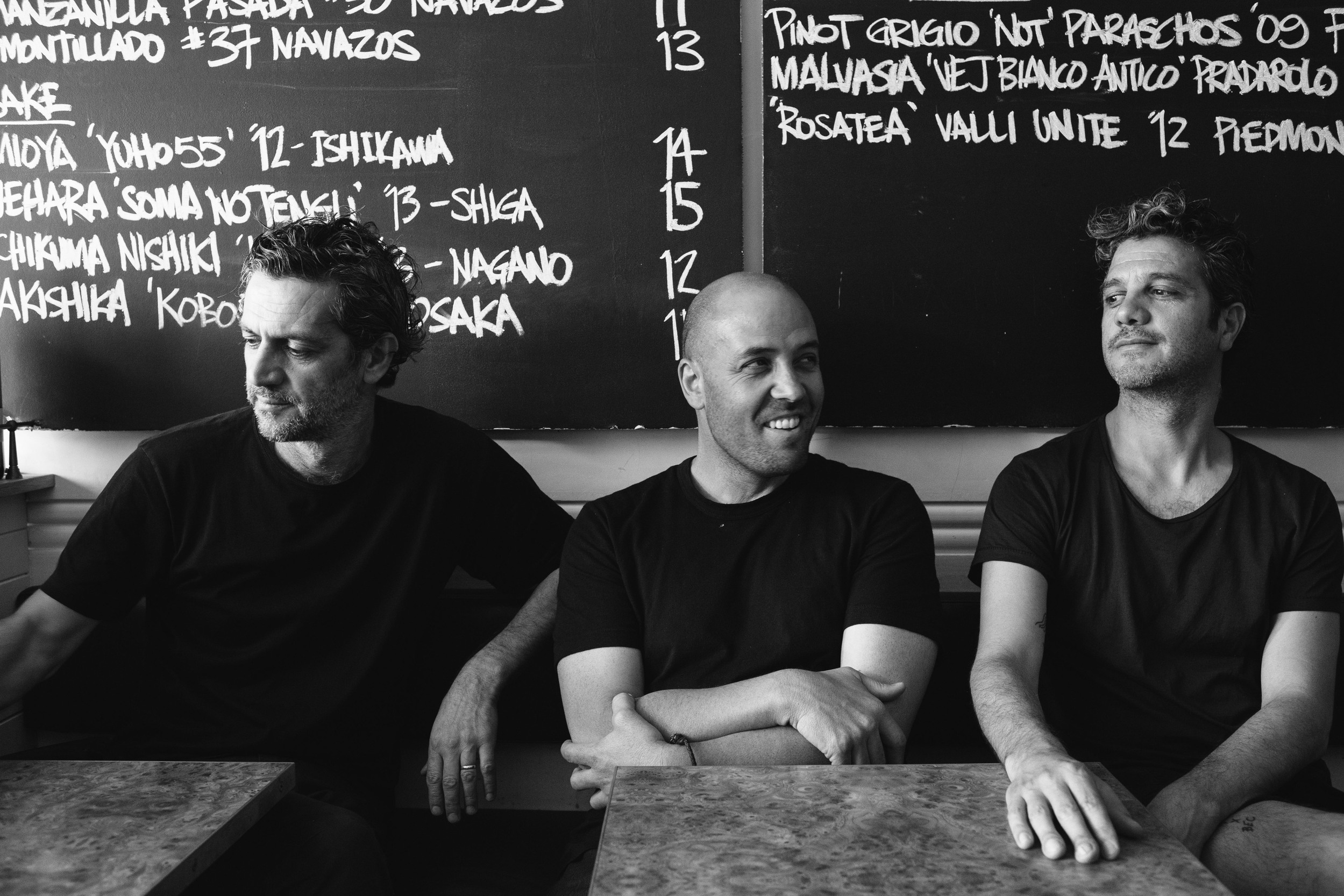 Black and white shot of three men sitting at a table in a restaurant, with a menu board visible in the background, indoor.