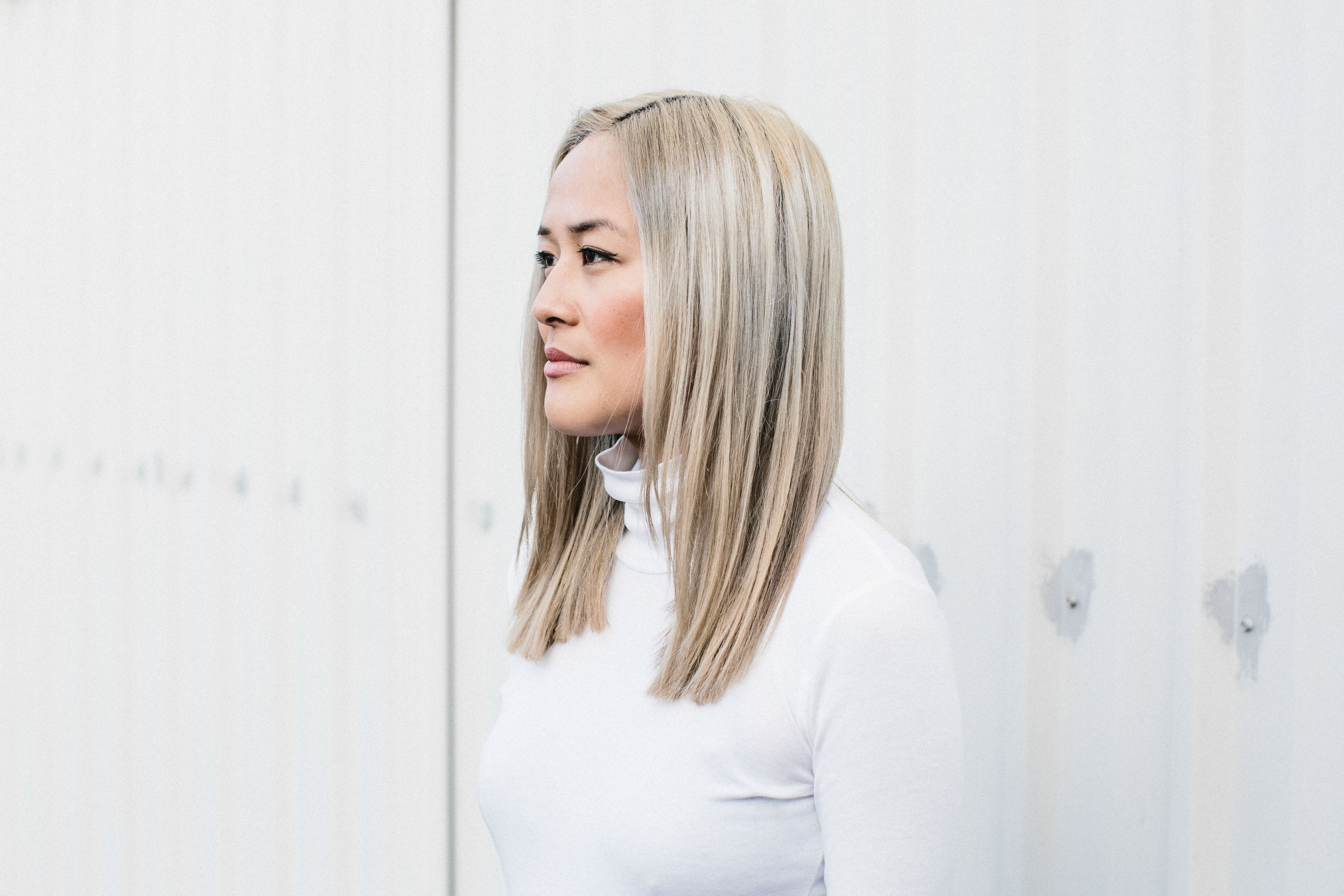 Profile portrait of a blonde woman in a white turtleneck against a plain white wall, showcasing a clean and minimalist aesthetic.