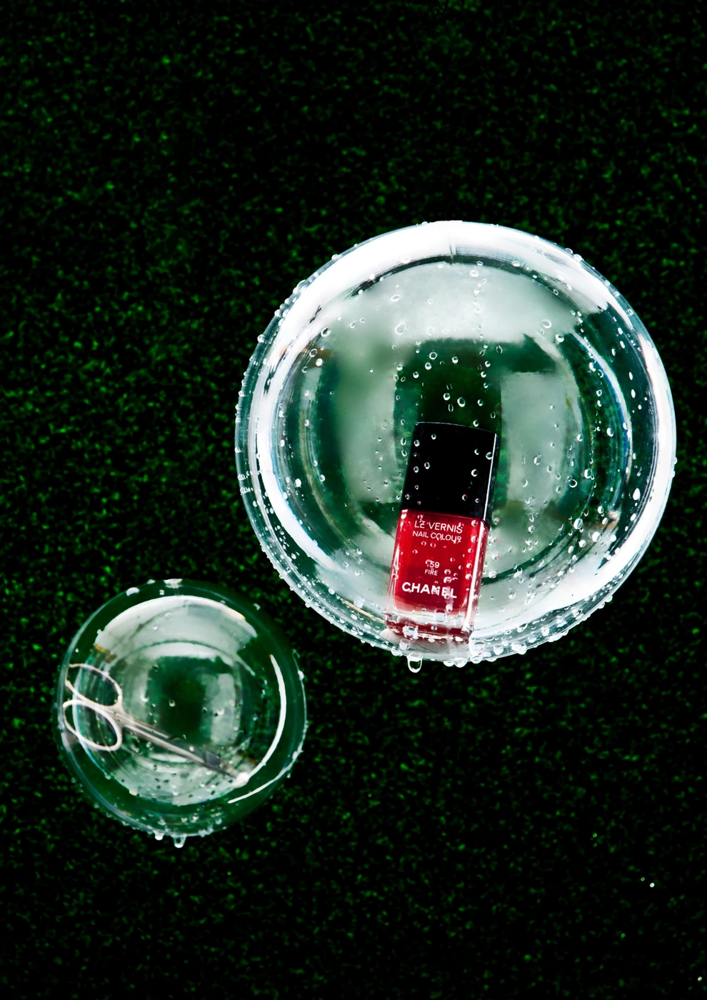 Overhead shot of a red nail polish bottle and scissors inside glass bowls on green textured turf