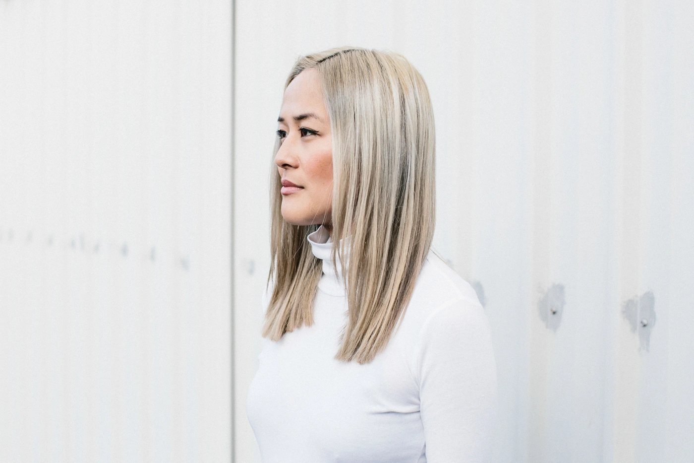 Portrait of an East Asian woman with straight blonde hair wearing a white turtleneck against a white wall.