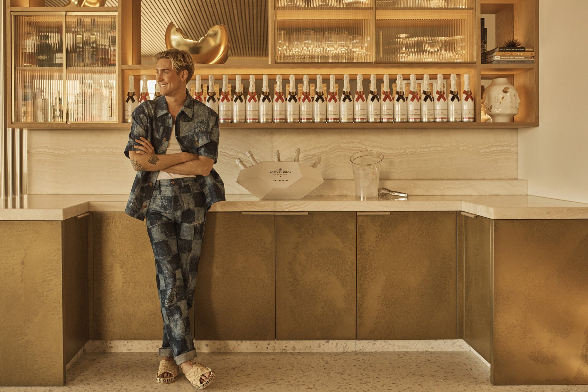 Stylish man in a patchwork suit stands in an interior with champagne bottles, gold cabinetry, and textured walls and floors.
