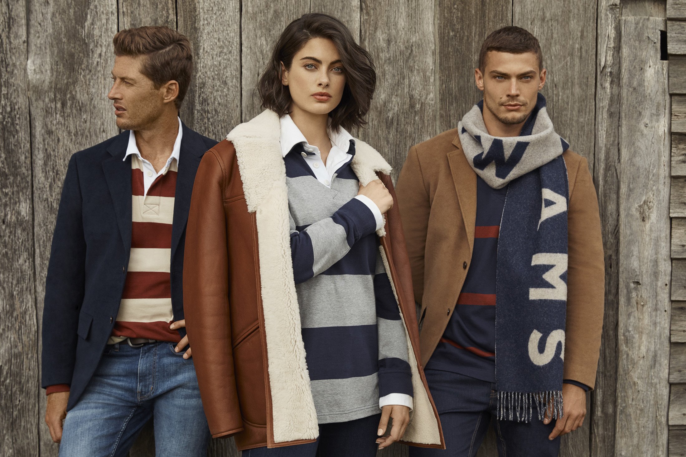 Three fashion models pose outdoors wearing striped shirts, jackets, and a scarf against a rustic wooden wall in a medium studio shot.