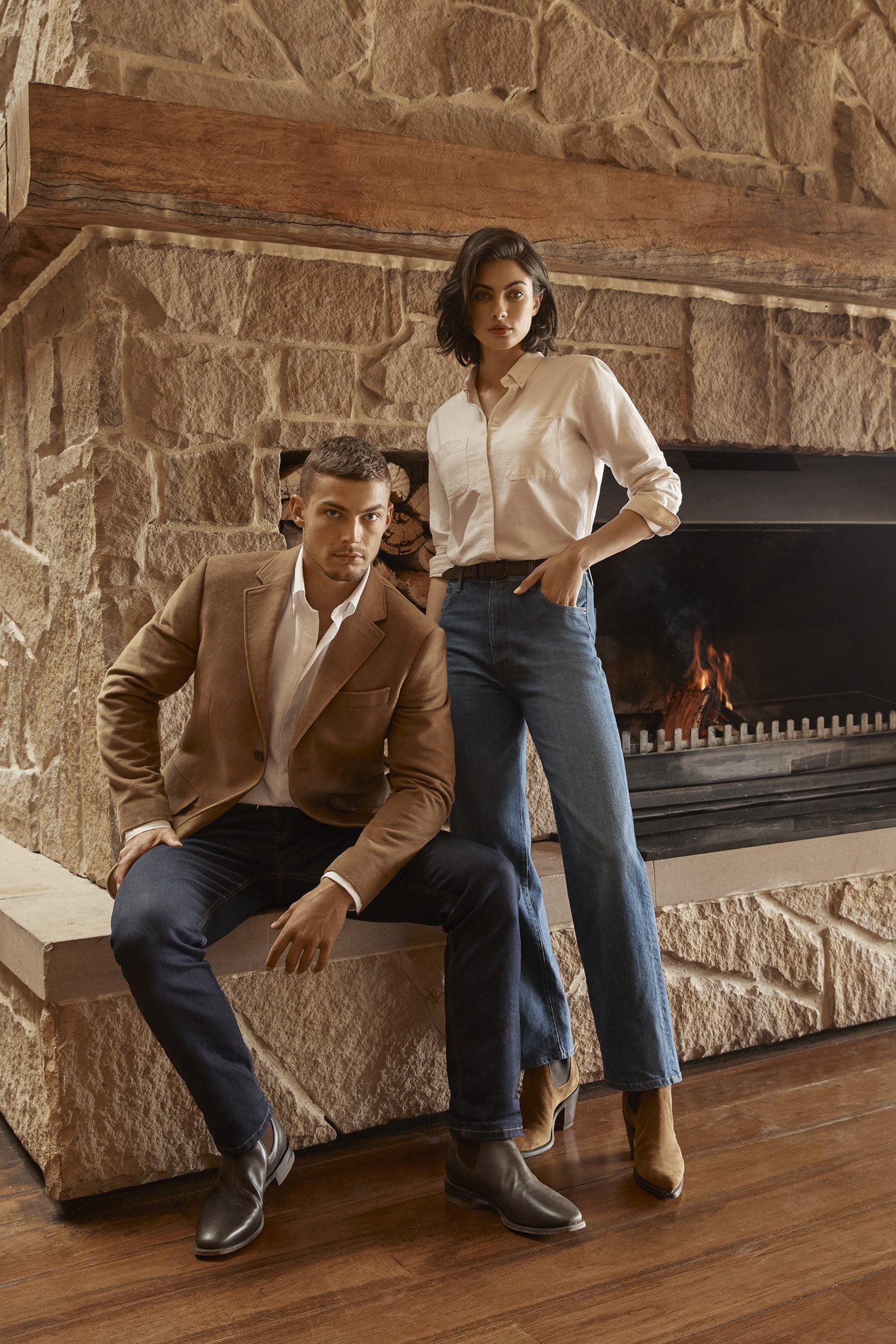 Man and woman pose in front of stone fireplace in stylish casual clothing, including a blazer, collared shirt, and jeans.