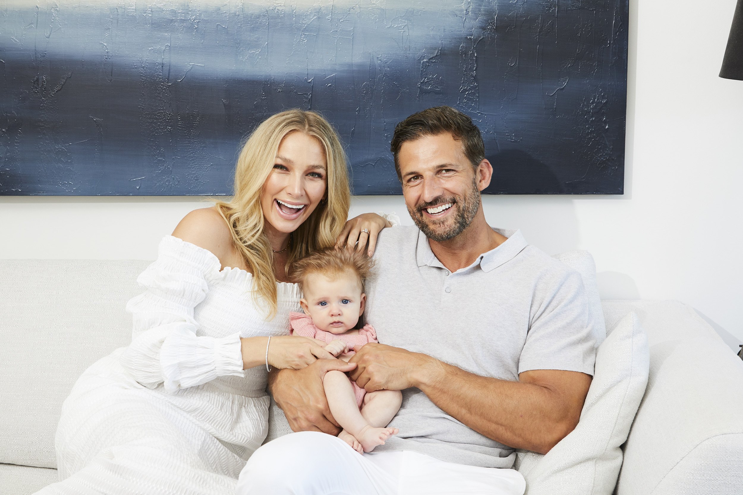 Smiling family of three on a white couch in a brightly lit living room with white walls, a baby, and an abstract painting.