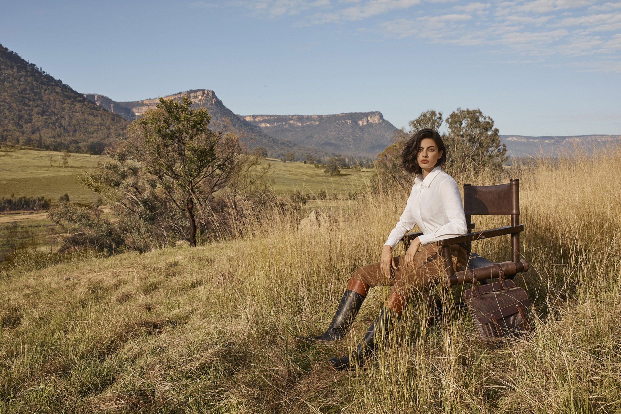 Woman in vintage riding attire poses on a chair in a grassy field with distant mountains, bathed in soft natural light.