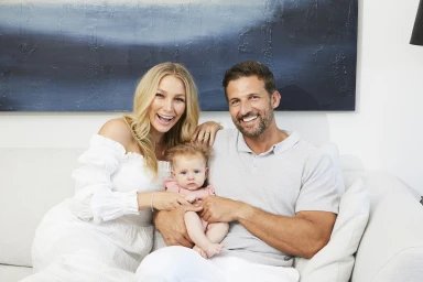 Smiling man and woman holding a baby sitting together on a light sofa against a blue abstract wall art background