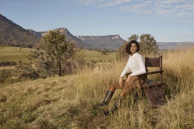 Woman in white shirt and riding boots seated in a leather director chair in a grassy valley with mountain backdrop