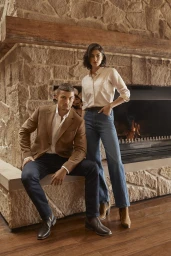 A man seated and a woman standing in front of a large stone fireplace styled by fashion stylist Pip Cook