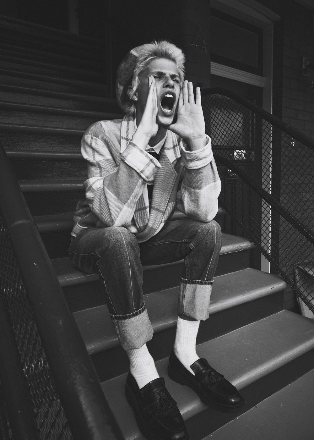 Monochrome portrait of a young adult on outdoor steps, calling out with hands cupped around their mouth, wearing casual plaid jacket and denim jeans.
