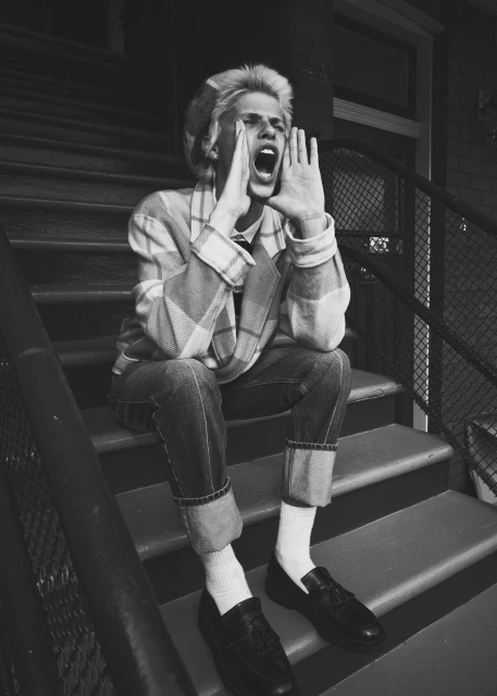 Monochrome portrait of a young adult on outdoor steps, calling out with hands cupped around their mouth, wearing casual plaid jacket and denim jeans.