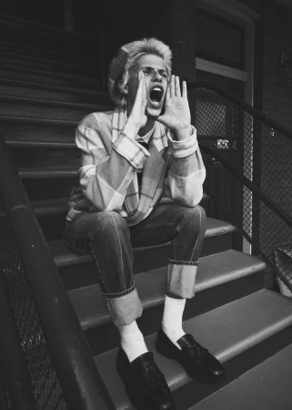 Monochrome portrait of a young adult on outdoor steps, calling out with hands cupped around their mouth, wearing casual plaid jacket and denim jeans.
