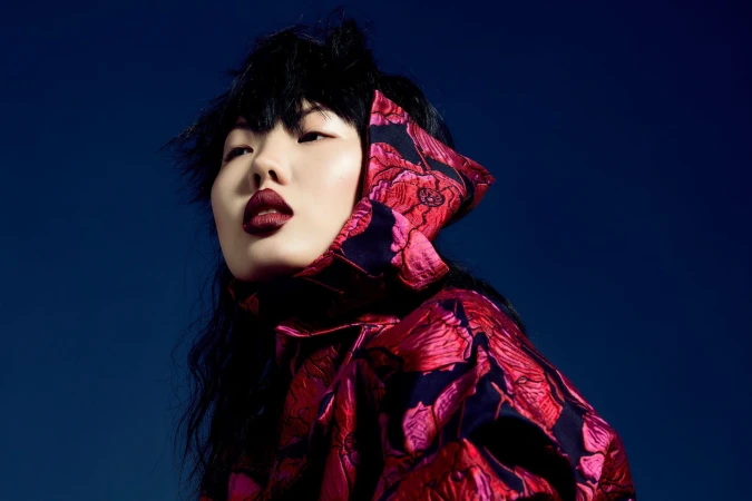 Close-up portrait of an adult woman with dark hair and bold dark red lipstick wearing a red floral-patterned hood against a blue background.