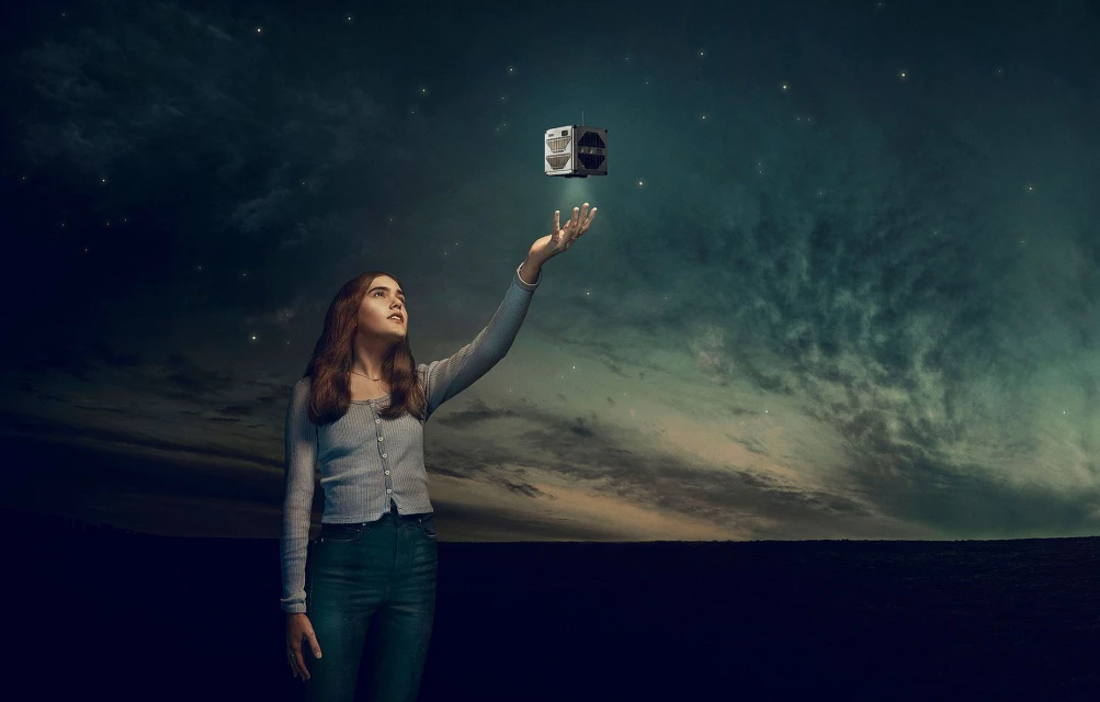 Young woman reaches toward a floating, cube-shaped object in a dark, dreamy landscape under a starry sky with soft, diffused lighting.