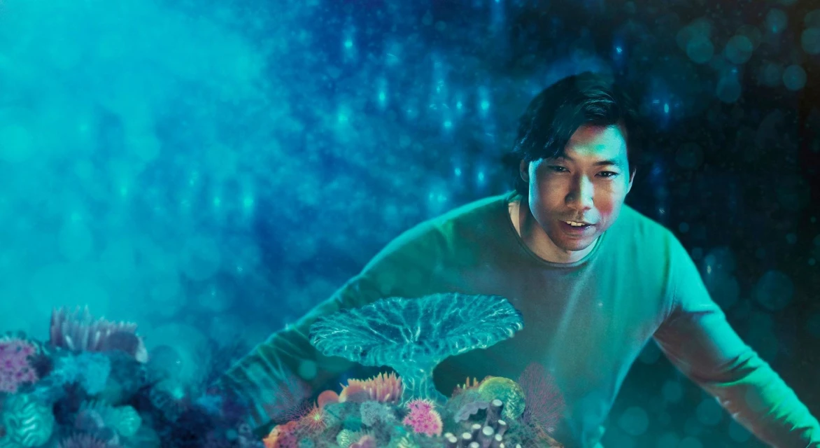 Man wearing green stands amongst coral reef against an abstract turquoise background, suggesting underwater environment and digital art.