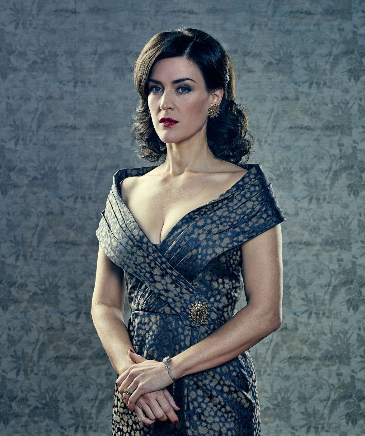 Portrait of a woman wearing an off-the-shoulder patterned dress and earrings against a patterned wallpaper background