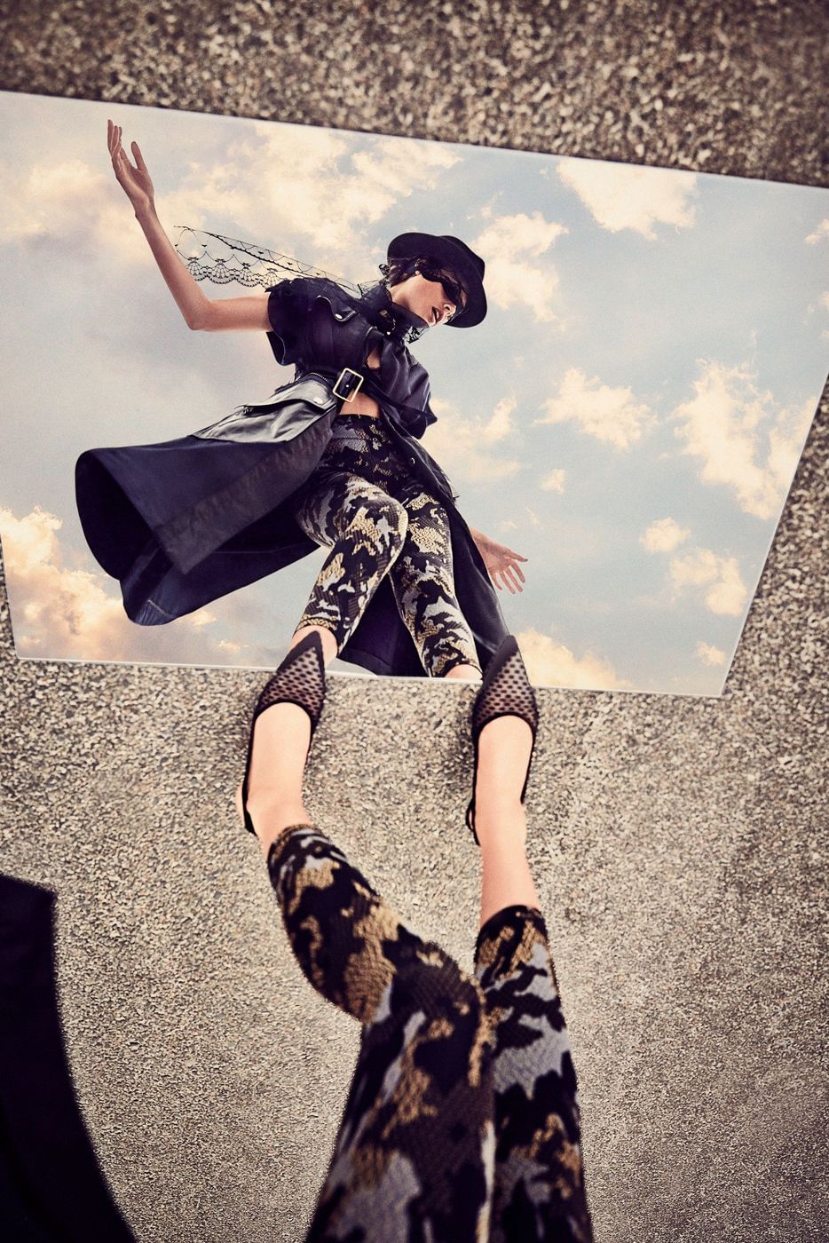 Fashion shoot featuring a model wearing a dark jacket, camo pants, black shoes, and a hat, reflected in a rectangular mirror.