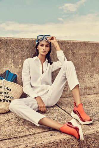 Woman in white linen outfit and red slip-on sneakers sitting on concrete ledge with beach bag and swim goggles on head.