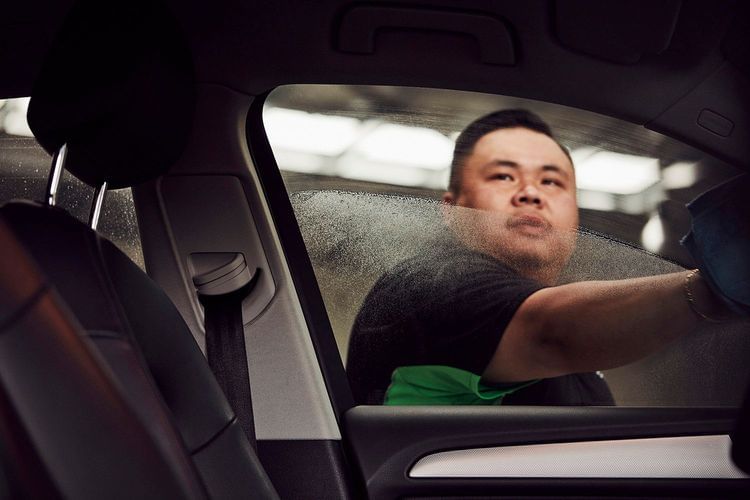 Adult Asian man cleaning the inside of a car window with a microfiber cloth, inside a car with black interior, medium shot.