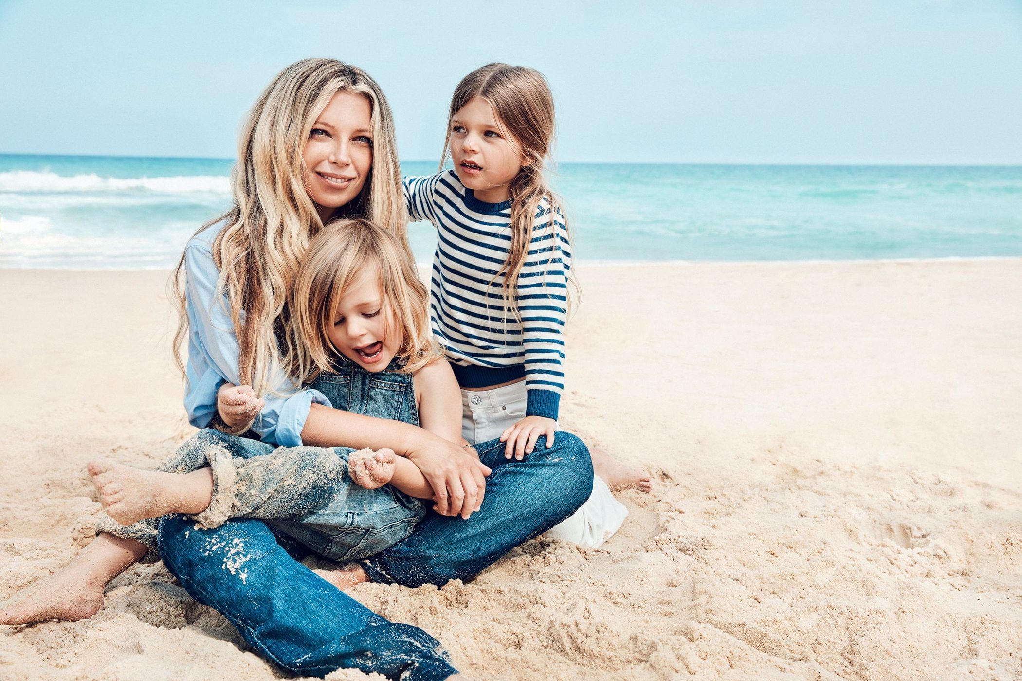 Blonde mother and two young daughters sitting on a sandy beach near turquoise ocean on a sunny day, one child in overalls, family.
