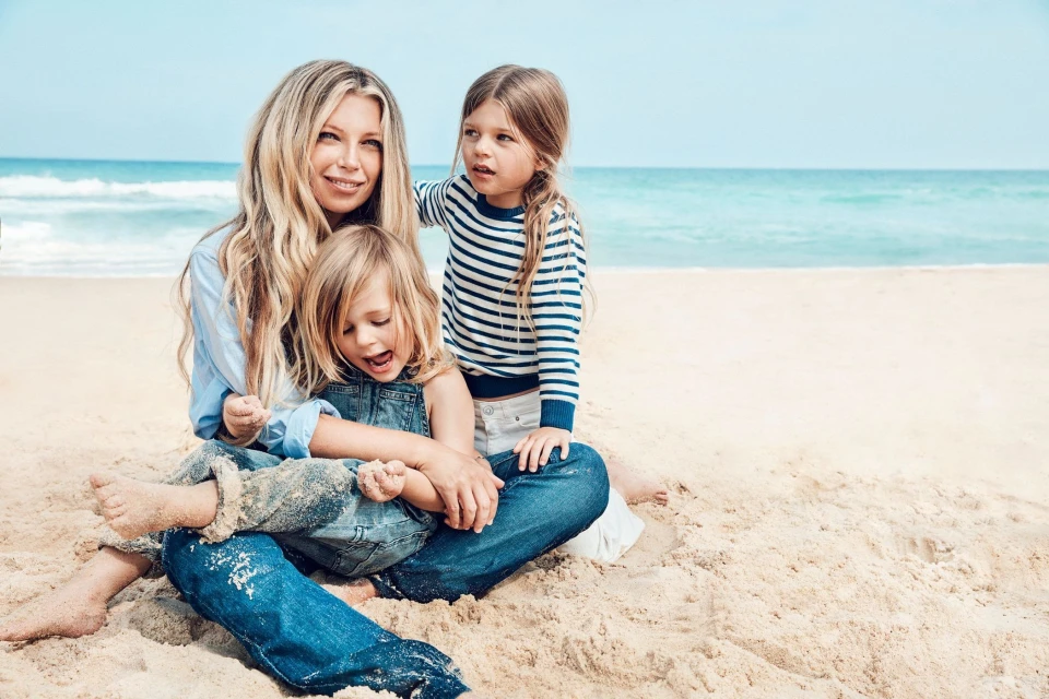Blonde mother and two young daughters sitting on a sandy beach near turquoise ocean on a sunny day, one child in overalls, family.