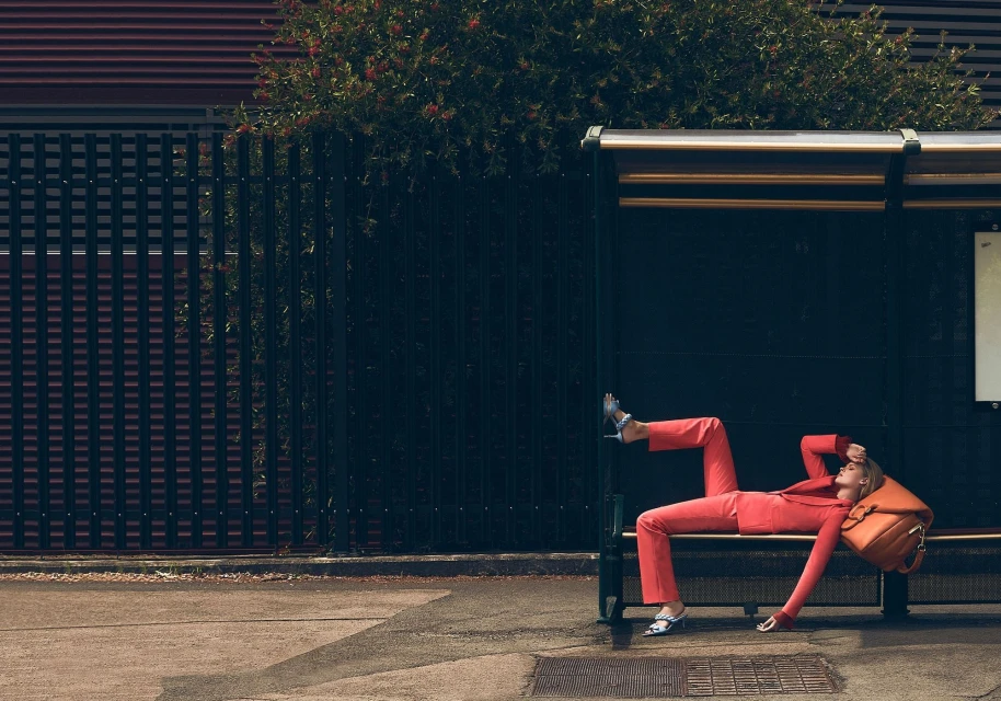 Woman in coral suit and heels lounging at bus stop with a backpack and foliage in the background. Outdoor shot with natural lighting.