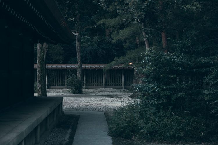 Exterior view of a traditional Japanese building and garden at night with gravel path, trees, and a covered walkway in muted, desaturated colors.