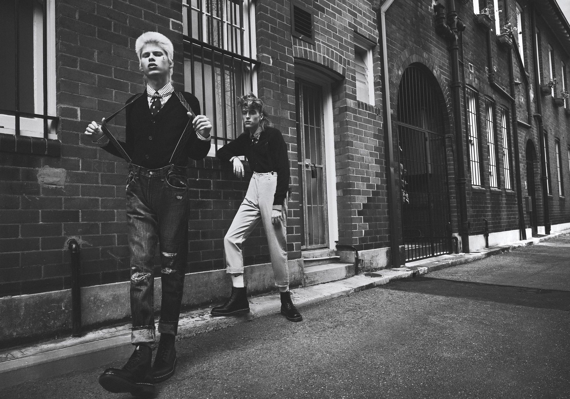 Two young adult fashion models in black and white stand outside a brick building in jeans, suspenders, and boots in an outdoor setting.